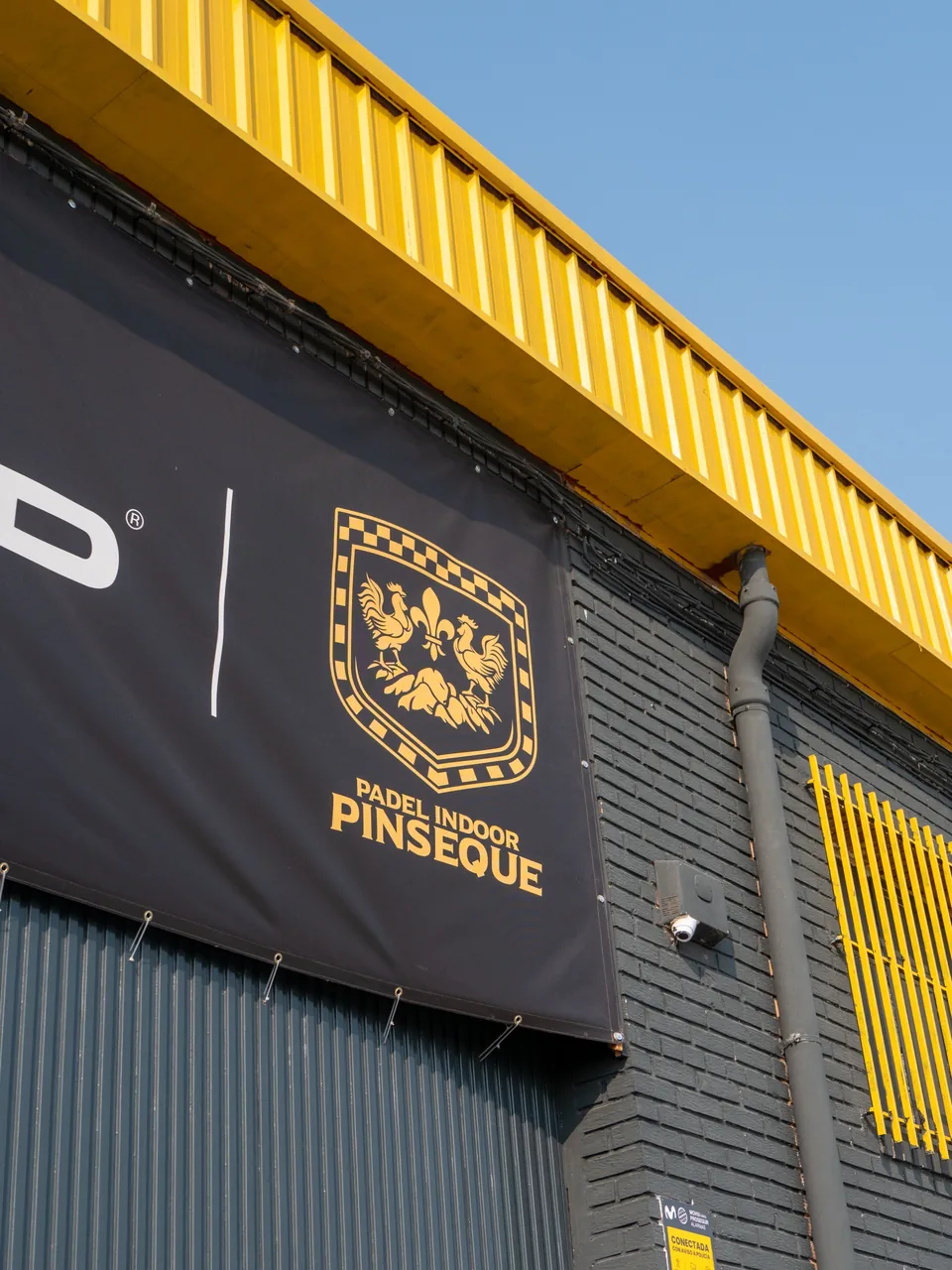 Fachada de Pádel Indoor Pinseque con el escudo del club y la cubierta amarilla